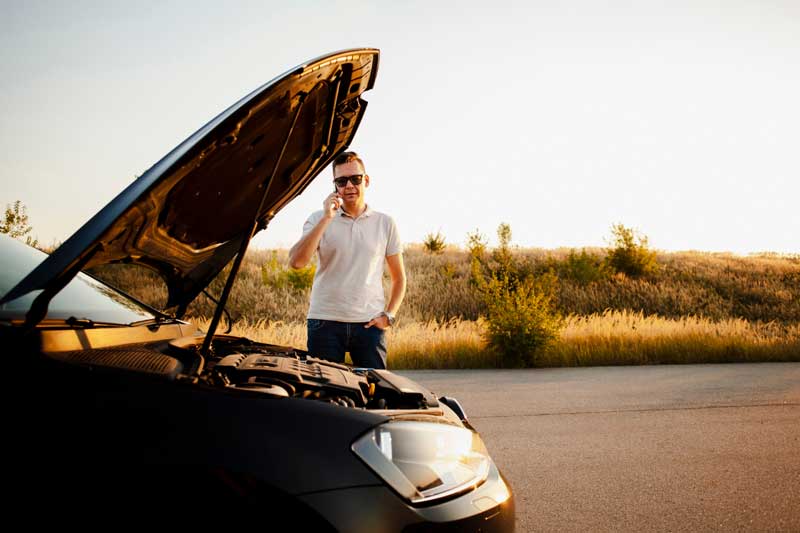 Eine Person in einem weißen Hemd steht neben einem Auto mit offener Motorhaube und spricht am Telefon vor einem Sonnenuntergangshorizont.