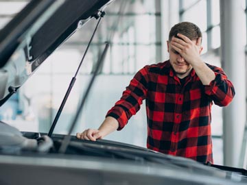 Eine Person in einem rot-schwarzen Flanellhemd inspiziert eine offene Motorhaube und zeigt Besorgnis in einer gut beleuchteten Garage.